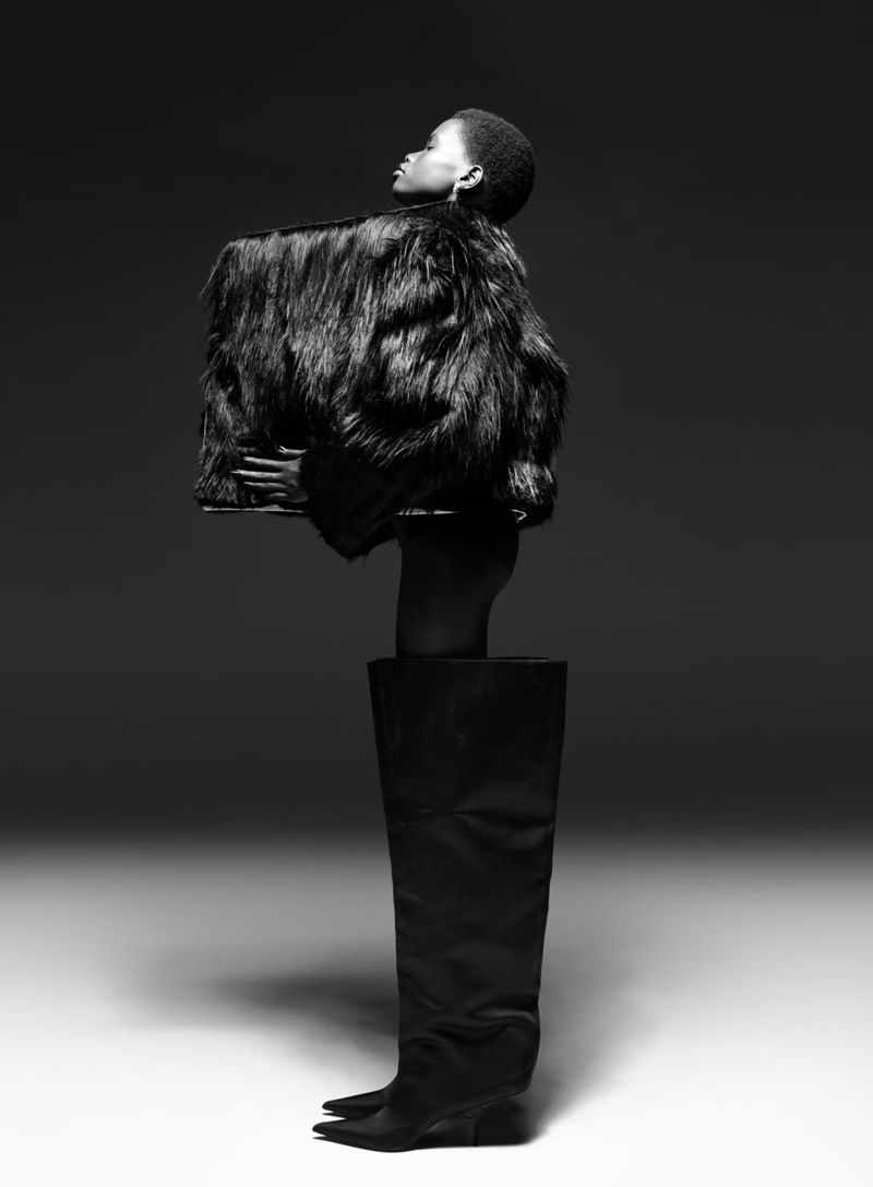 A person with short hair wearing a voluminous faux-fur jacket and one giant, oversized black boot stands against a plain background.