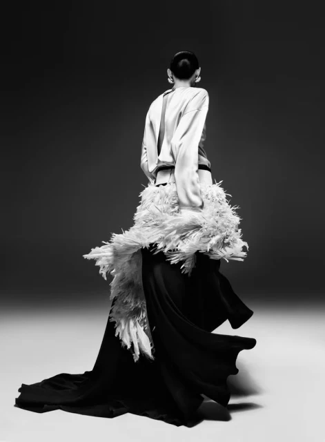 Back view of a woman in a long black skirt and white top with feathered accent, captured in a studio shot with diffused lighting.