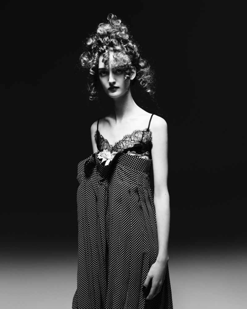 Black and white portrait of a woman with curly hair, wearing a lace top and polka-dot dress, standing in a photo studio setting.