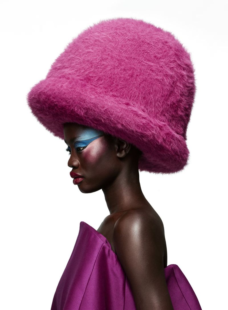 Fashion portrait of a woman with dark skin wearing a large pink furry hat and magenta garment, with creative blue eye makeup, side view.