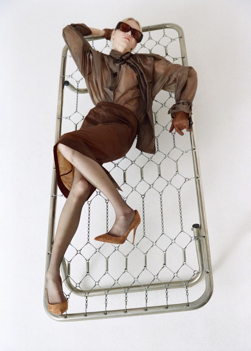 An adult female fashion model reclines on a metal frame in a studio, wearing a brown sheer blouse, skirt, heels, sunglasses, and gloves against a white background.