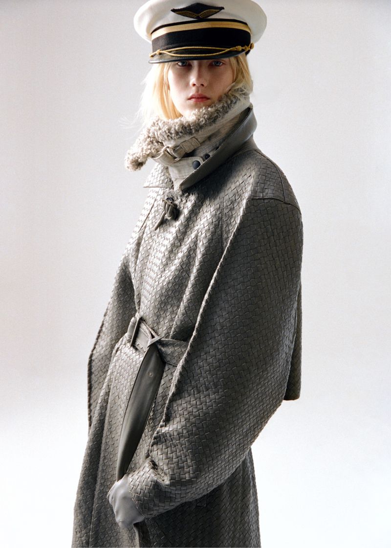 Woman in patterned gray coat and pilot cap poses in front of a neutral background in a fashion shot.