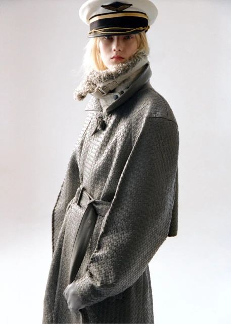 Woman in patterned gray coat and pilot cap poses in front of a neutral background in a fashion shot.