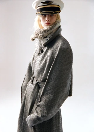 Woman in patterned gray coat and pilot cap poses in front of a neutral background in a fashion shot.