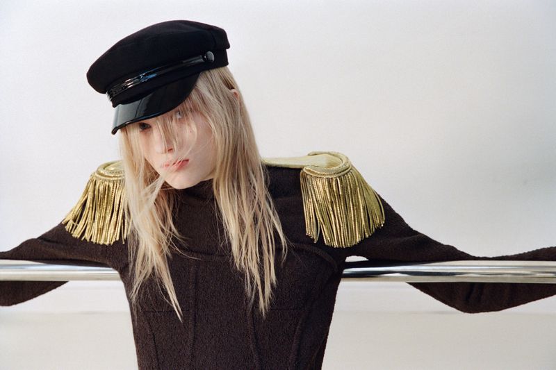 A model in a dark brown top, wearing a black cap and gold epaulettes, leans against a silver metal bar, set against a white backdrop.