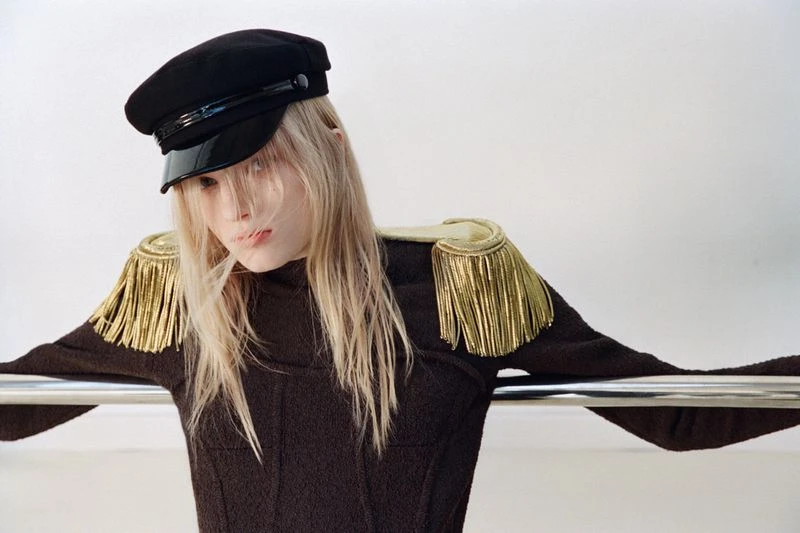 A model in a dark brown top, wearing a black cap and gold epaulettes, leans against a silver metal bar, set against a white backdrop.