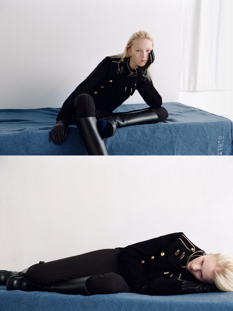 Model in black military-style jacket and tall boots resting on a blue blanket against a plain white wall