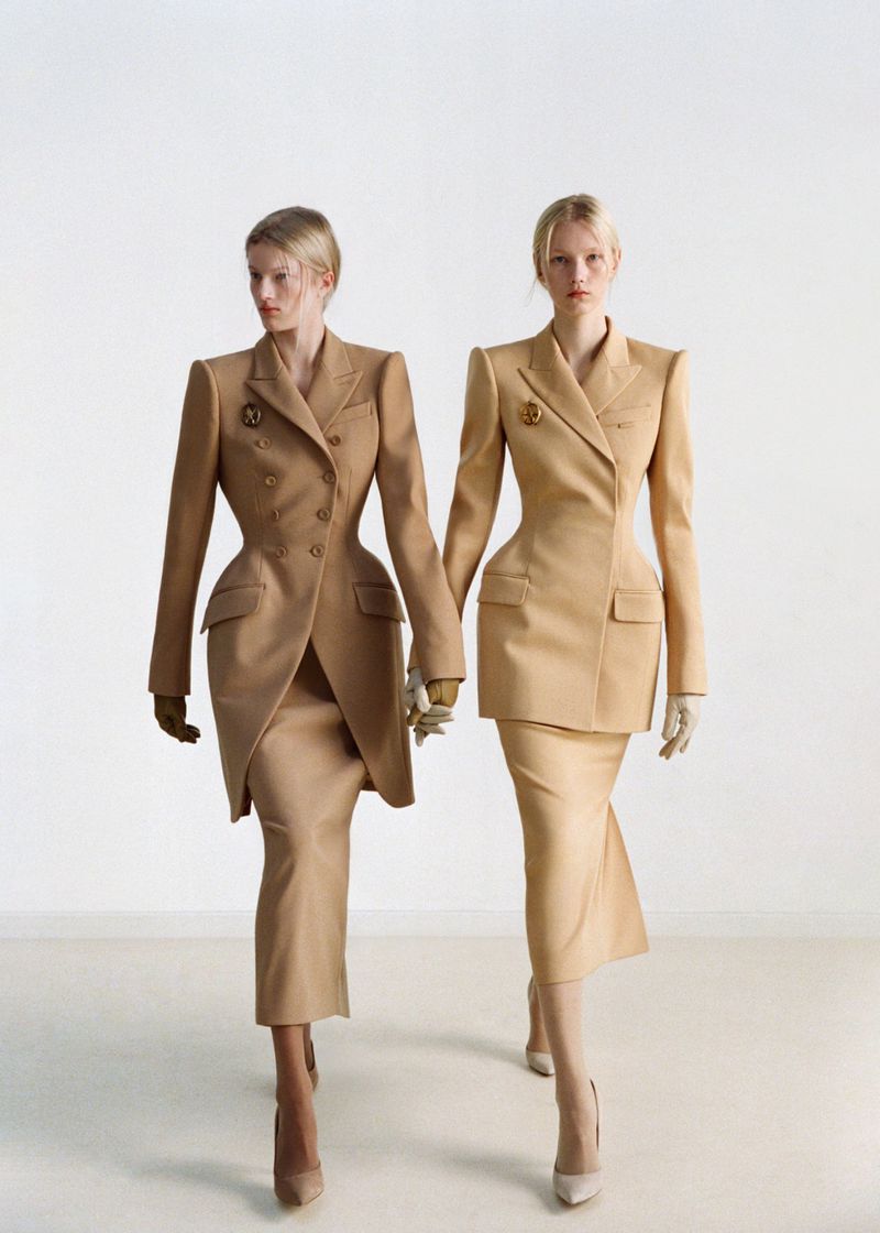 Two blonde women in beige skirt suits walk hand-in-hand against a white background in a fashion editorial style shot.