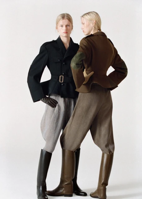 Two fair-skinned female models pose in high-fashion vintage-inspired jackets, jodhpurs, and riding boots against a white backdrop in a studio setting.