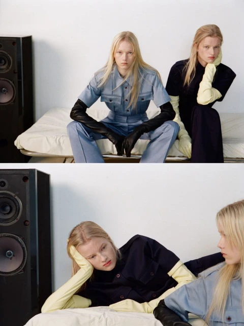 Two young fair-skinned women sitting side-by-side, in a studio, wearing matching workwear-inspired outfits and gloves, next to a speaker.