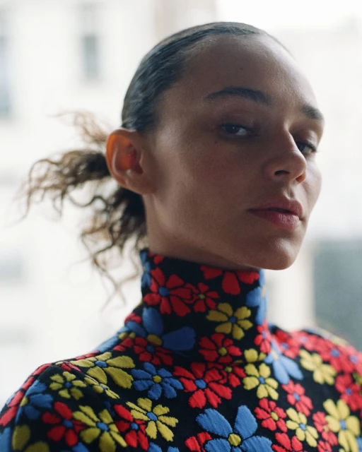 Close-up of a woman with dark hair styled back, wearing a floral print, high-necked top, in a bright, naturally lit indoor setting.