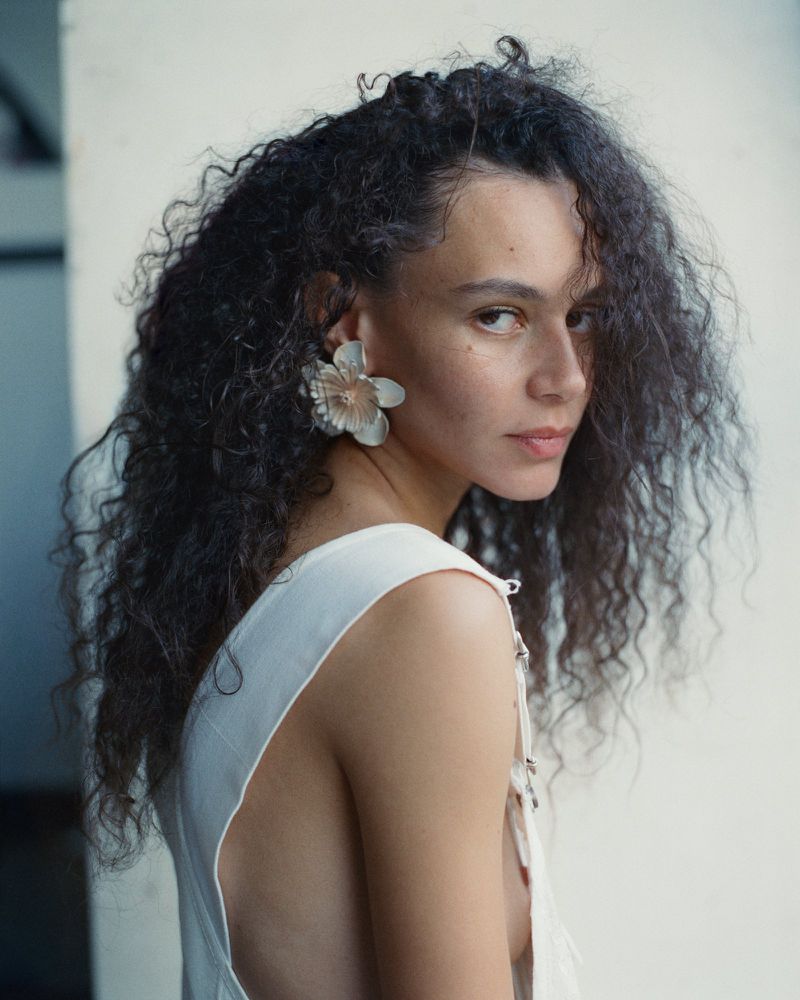 Portrait of a woman with curly hair wearing a white top and large flower earrings against a neutral backdrop. Beauty and fashion shot.