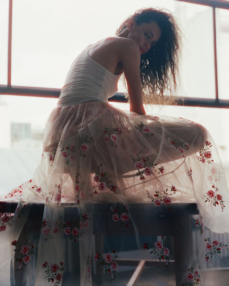 Woman in floral dress posing on a table in front of a window, soft light, fashion editorial, indoor setting.