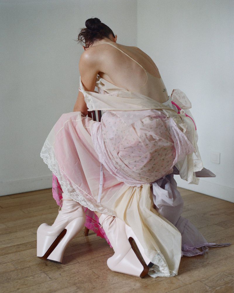 Back view of a person wearing an assortment of dresses, seated on a chair and wearing platform boots, against a minimal backdrop.