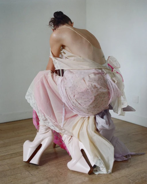 Back view of a person wearing an assortment of dresses, seated on a chair and wearing platform boots, against a minimal backdrop.
