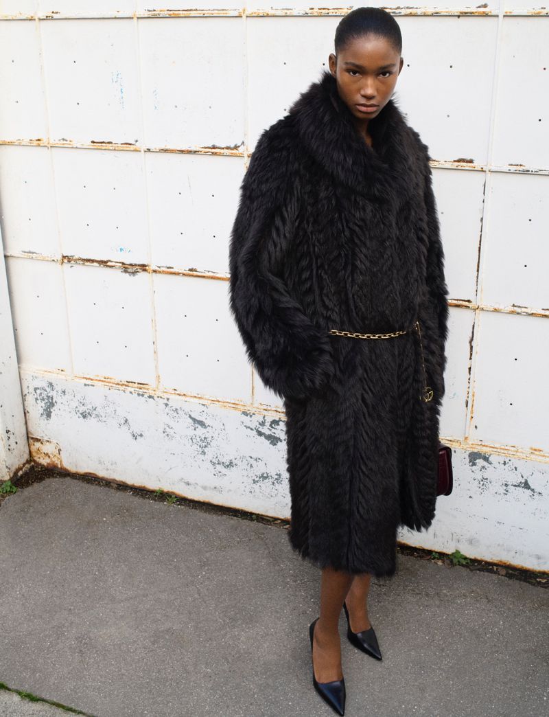 An African-descent female model poses in a long, dark fur coat with a gold chain belt and black heels against a distressed white wall.