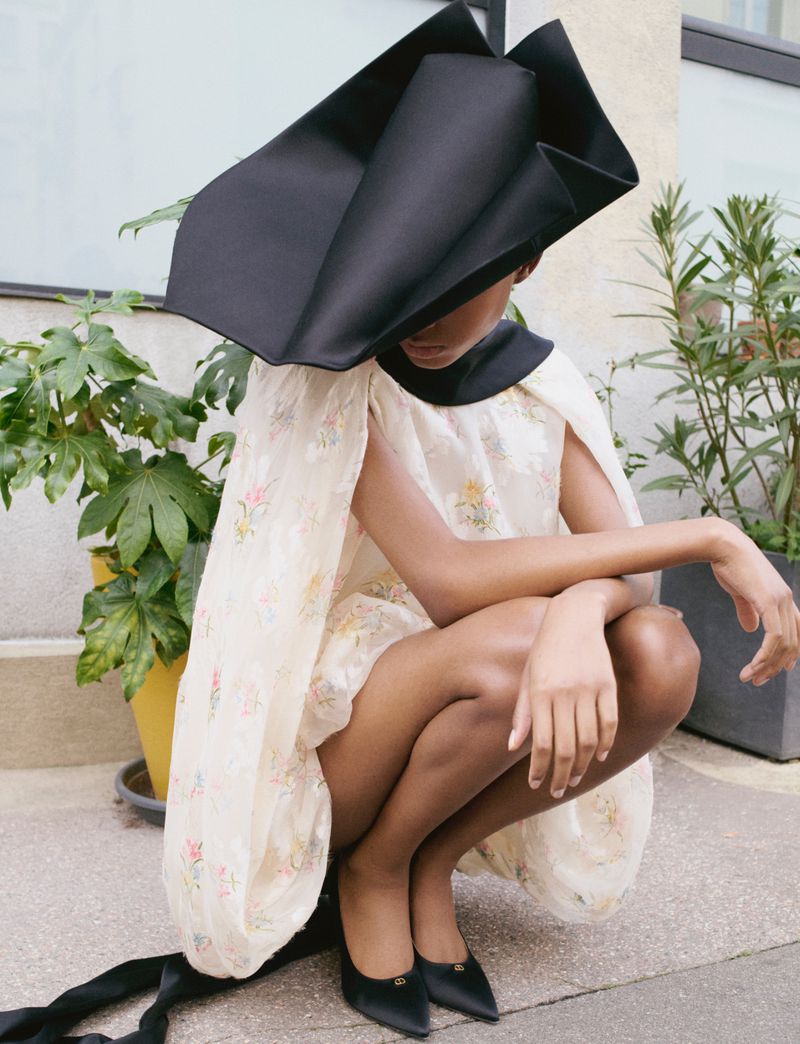 Fashionable woman crouches outdoors wearing floral dress, black pumps, and oversized black hat in a fashion editorial style.