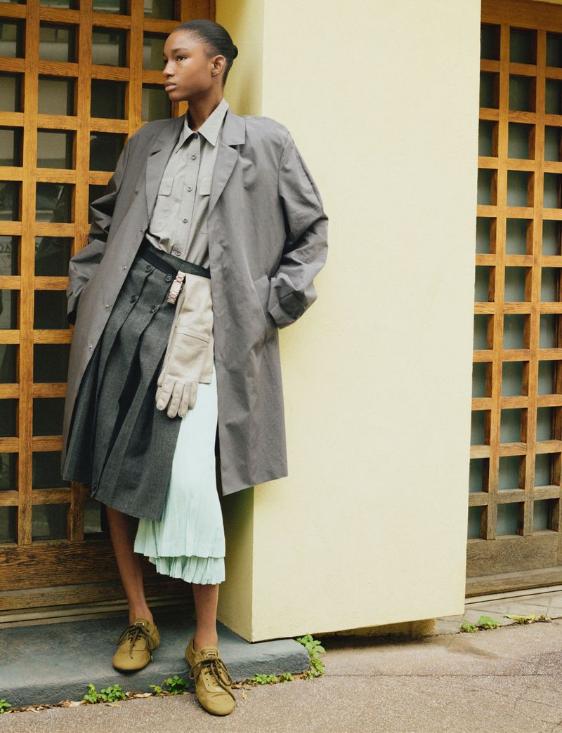Young woman in a grey trench coat over a pleated skirt posing outdoors against a geometric patterned wall, looking away.