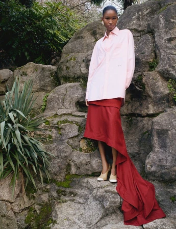 Model standing on rocky terrain wearing a light pink button-down shirt and a flowing red skirt with a long train.