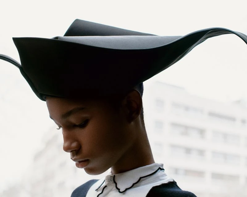 Close-up of Black woman in statement black hat with white-collared shirt, against a city apartment building backdrop.