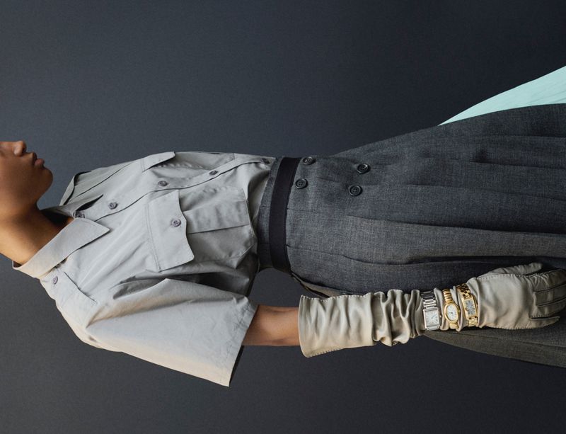 Fashion editorial shot of an adult model in a grey shirt and pants, accessorized with a belt, gloves, and multiple wristwatches, against a grey background.