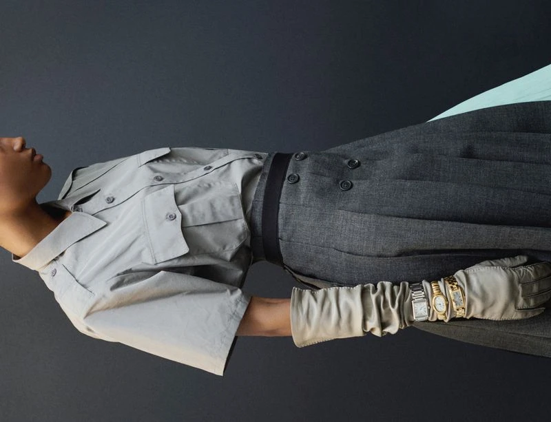Fashion editorial shot of an adult model in a grey shirt and pants, accessorized with a belt, gloves, and multiple wristwatches, against a grey background.