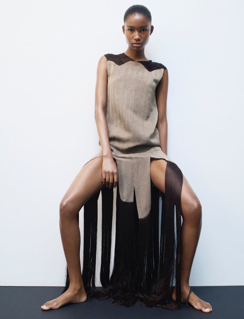 Striking fashion shot of a slender Black woman in a tan sleeveless dress with brown fringed skirt posing in a photo studio.
