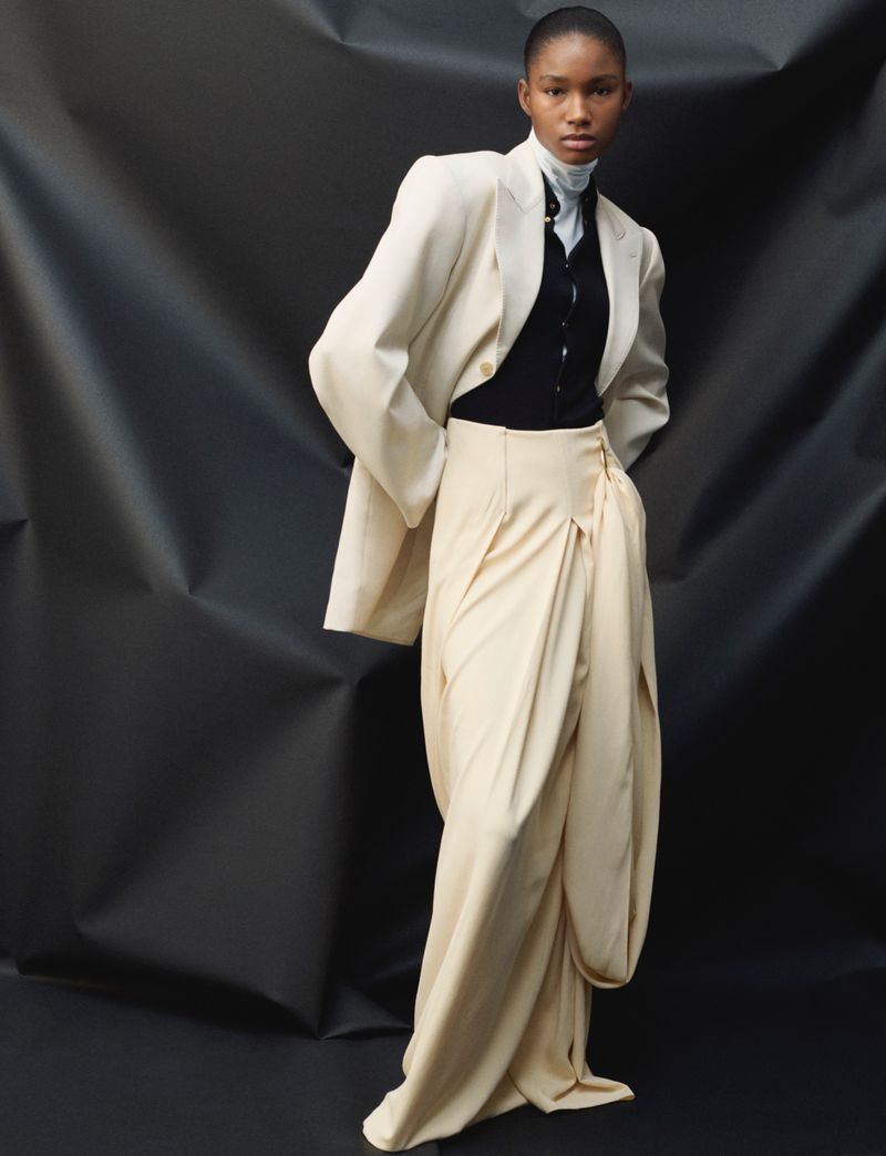Editorial fashion shot of a Black woman wearing an oversized cream-colored blazer with tailored pants in front of a dark, draped backdrop.