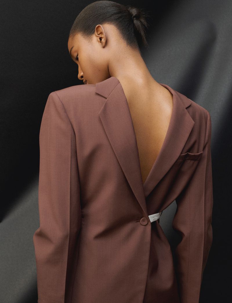 Woman in a brown blazer with a backless design poses against a dark background with soft lighting. The focus is on fashion and style.