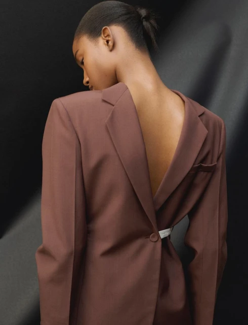 Woman in a brown blazer with a backless design poses against a dark background with soft lighting. The focus is on fashion and style.