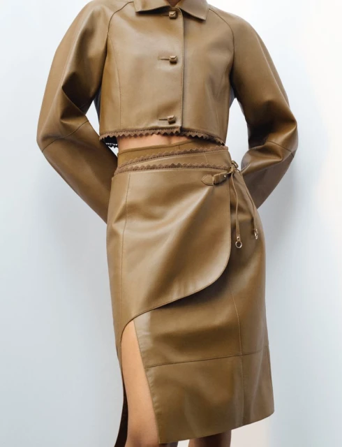 Woman wearing a tan, long-sleeved leather crop jacket and matching high-waisted skirt with a slit against a neutral backdrop.