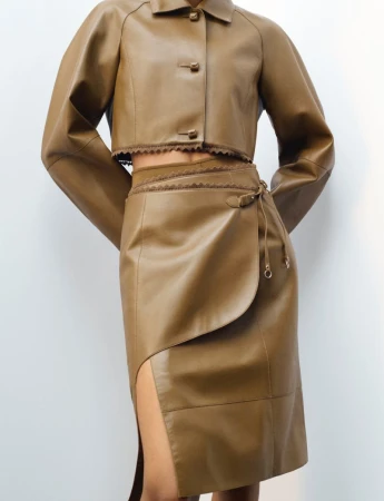 Woman wearing a tan, long-sleeved leather crop jacket and matching high-waisted skirt with a slit against a neutral backdrop.