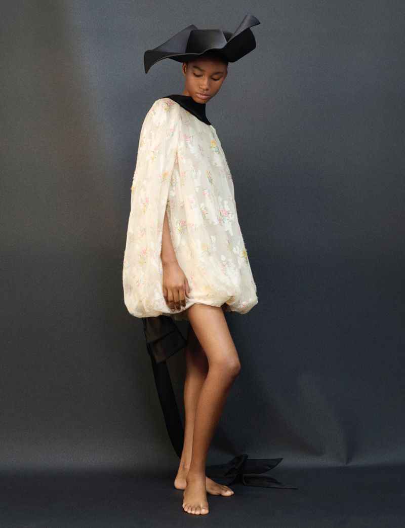 A slender Black fashion model poses in a studio wearing an ornate dress and a large brimmed hat against a gray backdrop in a fashion editorial.