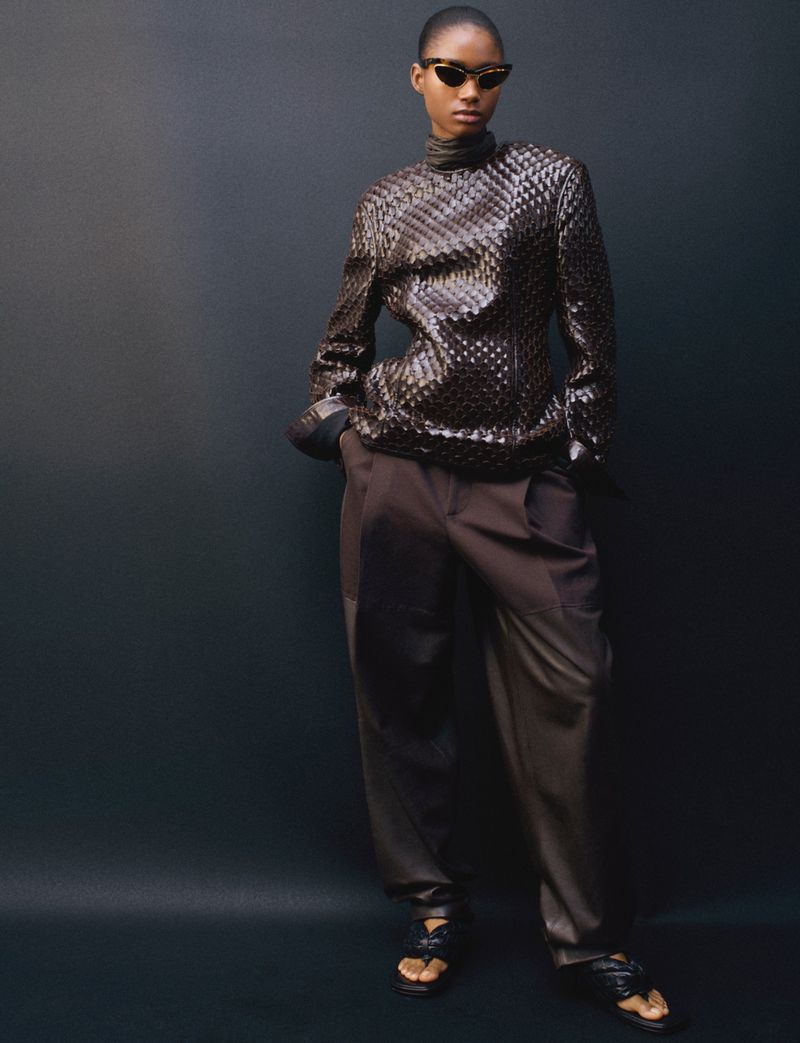 Elegant Black woman in a woven top and brown slacks poses in front of a dark backdrop wearing vintage-style sunglasses and strappy sandals.