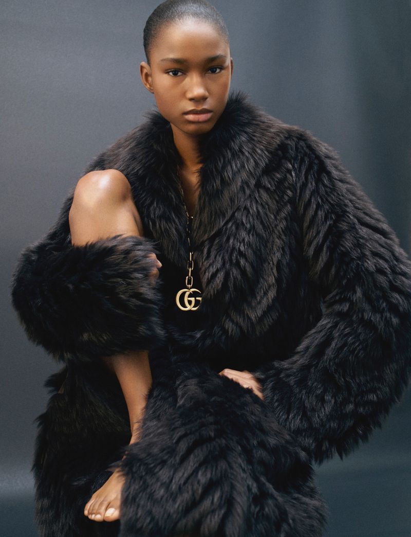 A black female fashion model poses in a dark faux fur coat with bare legs, and bare feet in a studio setting with a grey background.