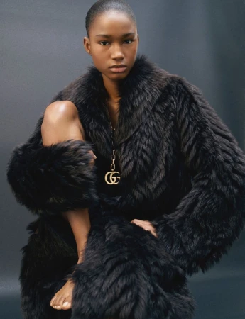 A black female fashion model poses in a dark faux fur coat with bare legs, and bare feet in a studio setting with a grey background.