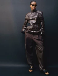 Model wearing dark cat-eye sunglasses, a textured brown long-sleeve top, and trousers against a dark backdrop