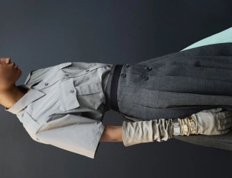 Model wearing a grey short-sleeve shirt and pleated skirt with layered watches over gloves against a dark backdrop.