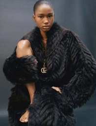 Model wearing a black furry coat and gold chain necklace against a dark grey studio background.