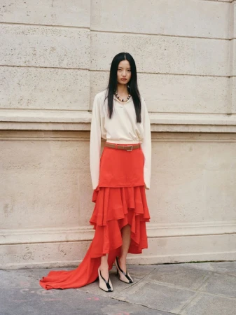 Model Chen Yang standing outdoors in a white top and tiered red skirt against a stone building wall for Muse Magazine by Sasha Marro.