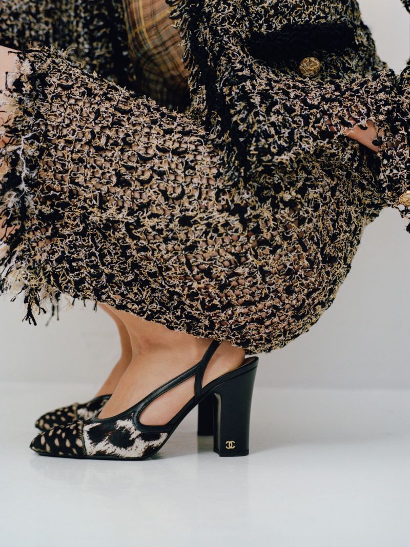 Black and beige tweed heels with a logo, worn with a tweed jacket and skirt on a white studio background, fashion editorial shot.