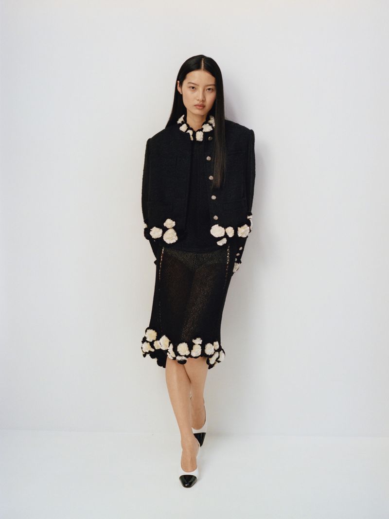 An Asian woman poses in a fashionable black floral-trimmed jacket and skirt with monochrome high-heeled shoes against a white backdrop.