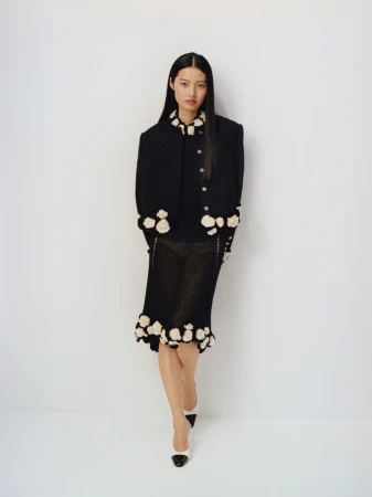 An Asian woman poses in a fashionable black floral-trimmed jacket and skirt with monochrome high-heeled shoes against a white backdrop.
