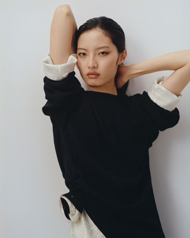 Portrait of an Asian female model with her arms raised behind her head, posing indoors against a clean white wall backdrop.