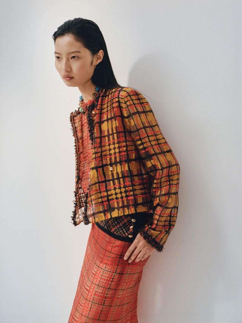 Asian woman poses in a patterned orange and black jacket and skirt against a plain white wall in a fashion editorial shot.