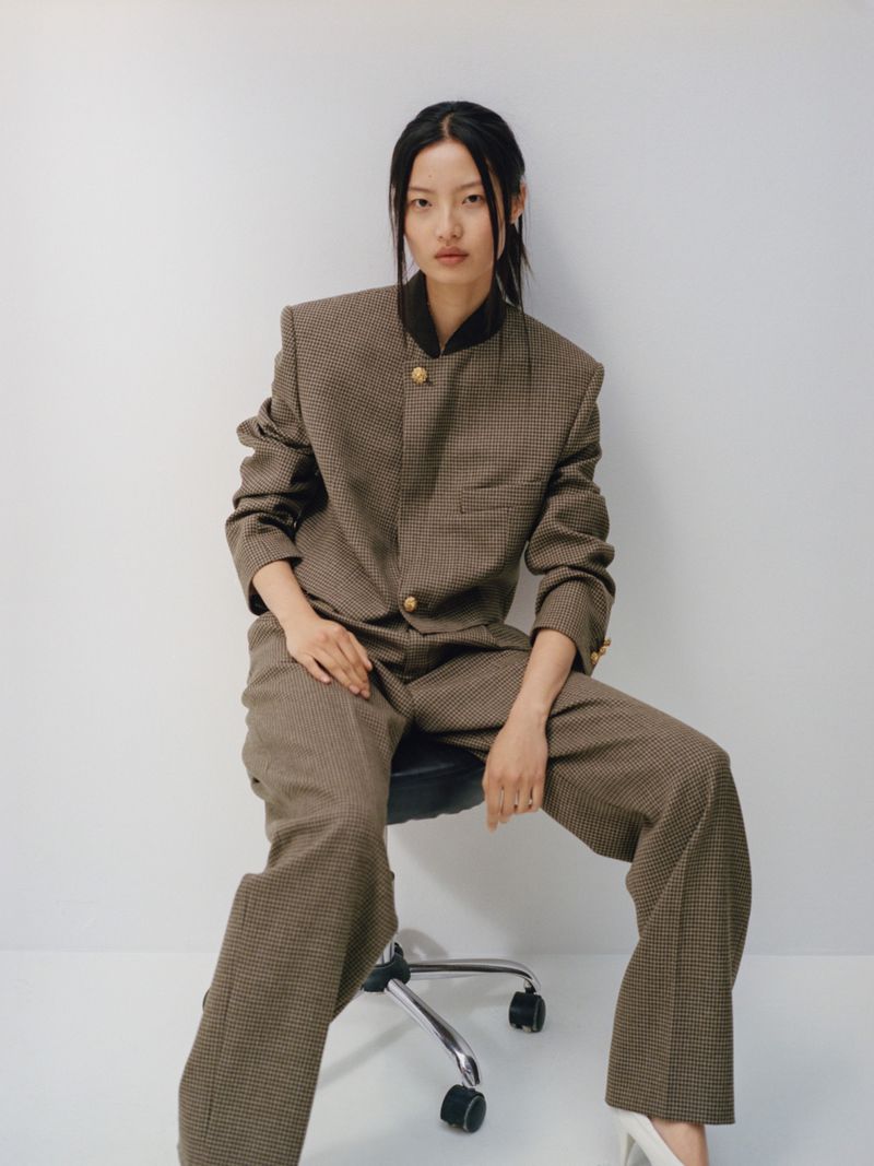 Elegant woman in a brown houndstooth suit seated on a rolling chair against a white background, gazing directly at the camera.
