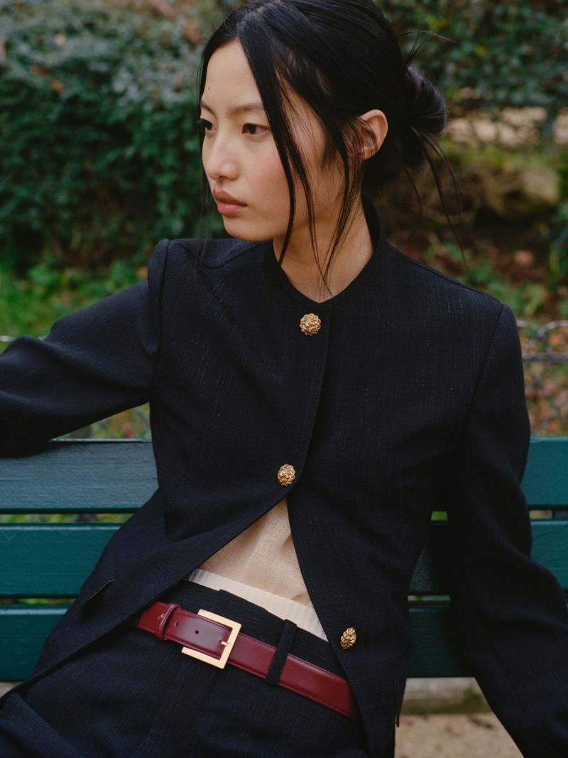 Portrait of a young Asian woman in a stylish black blazer with gold buttons, sitting outdoors on a park bench in soft, natural light.