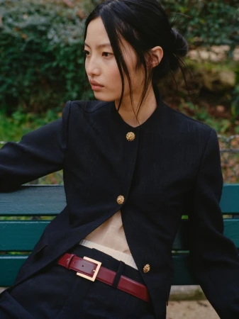 Portrait of a young Asian woman in a stylish black blazer with gold buttons, sitting outdoors on a park bench in soft, natural light.
