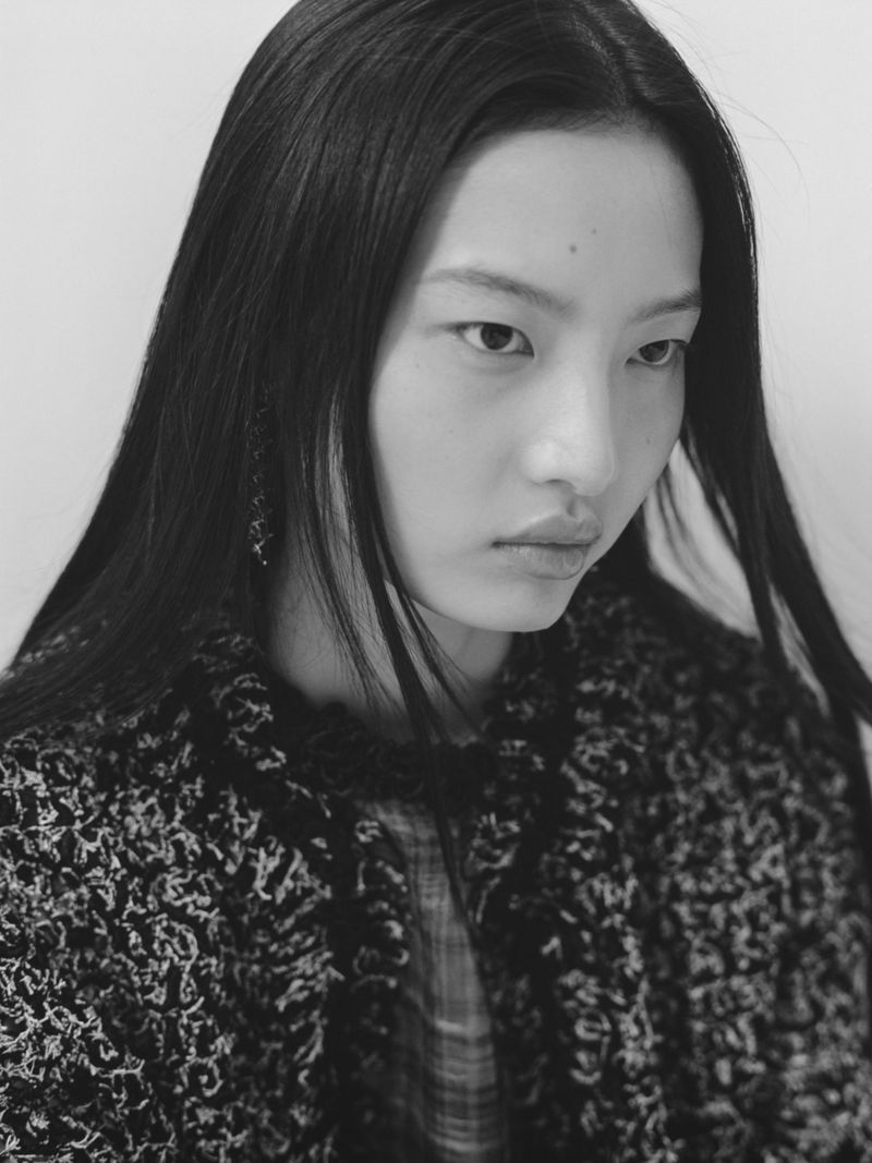 Close-up black and white portrait of an Asian woman with long dark hair wearing a textured black jacket looking off to the side.