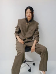 Model Chen Yang in a structured brown tweed suit seated on a rolling office chair against a clean white backdrop.